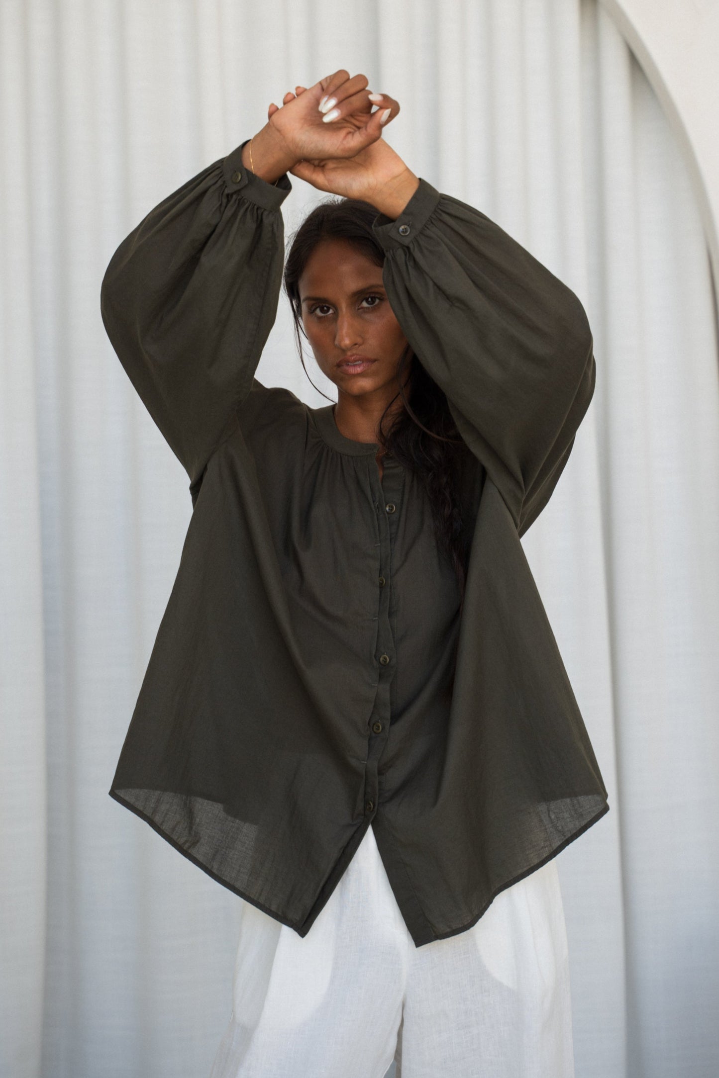 LILLY PILLY woman wearing a khaki Ember cotton voile shirt and ivory linen pants against a white curtain background.