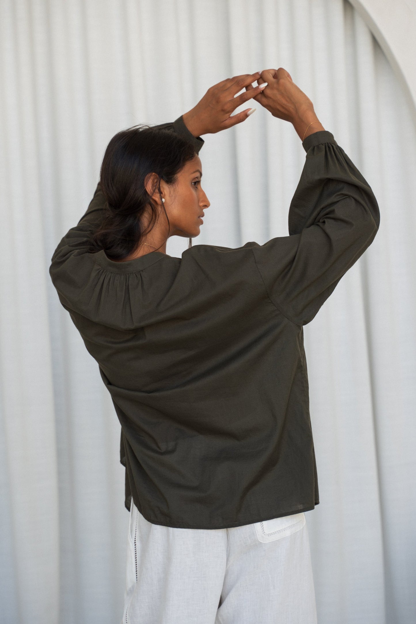 LILLY PILLY woman wearing a khaki Ember cotton voile shirt and ivory linen pants against a light curtain showing back view