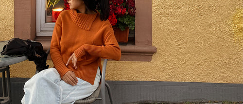 Woman in an orange sweater sitting on a chair against a yellow wall with a window and plants wearing Kaida knit in Cajun.