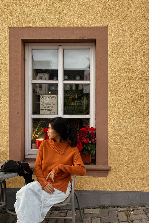 Woman in an orange sweater sitting on a chair against a yellow wall with a window and plants wearing Kaida knit in Cajun.