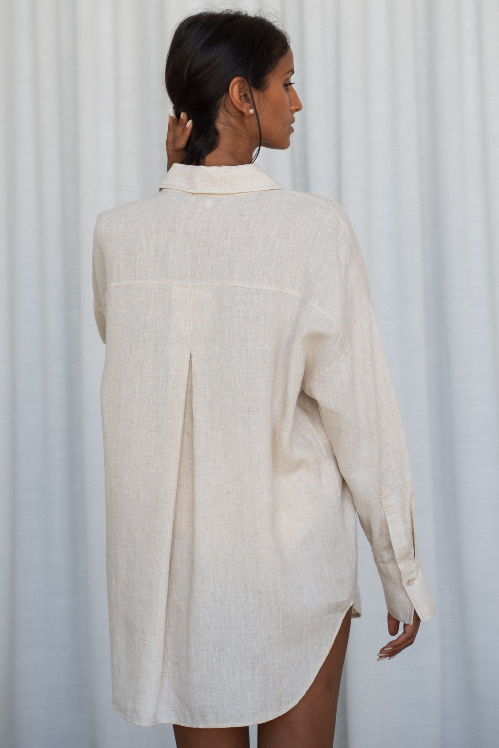 Woman wearing a oatmeal Kirra linen shirt, back view, against a white curtain background