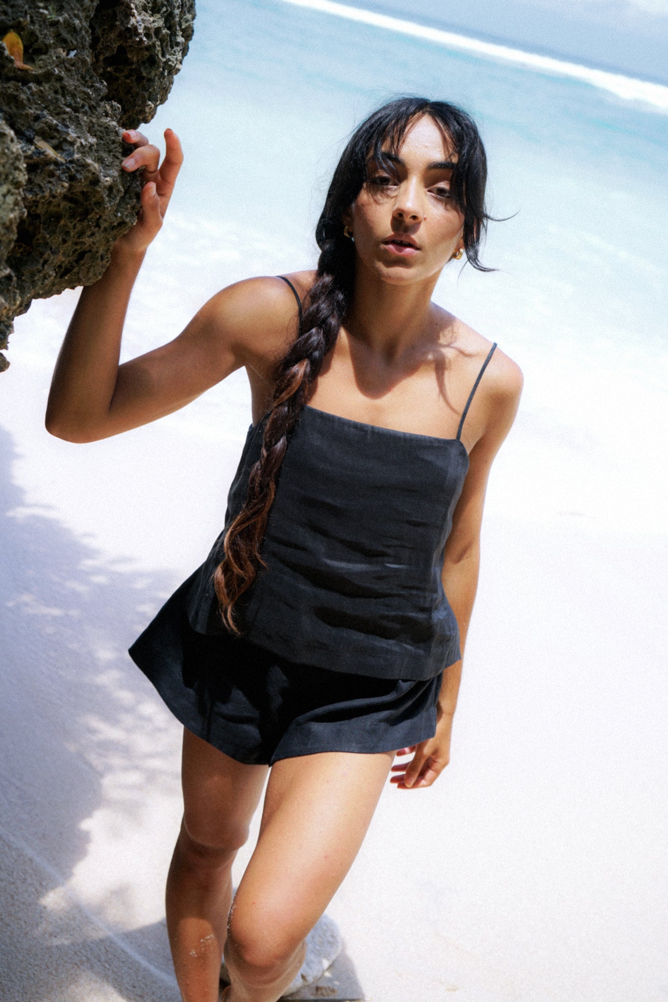 LILLY PILLY woman wearing a black Rosa silk linen cami top and shorts standing by a rock on a beach