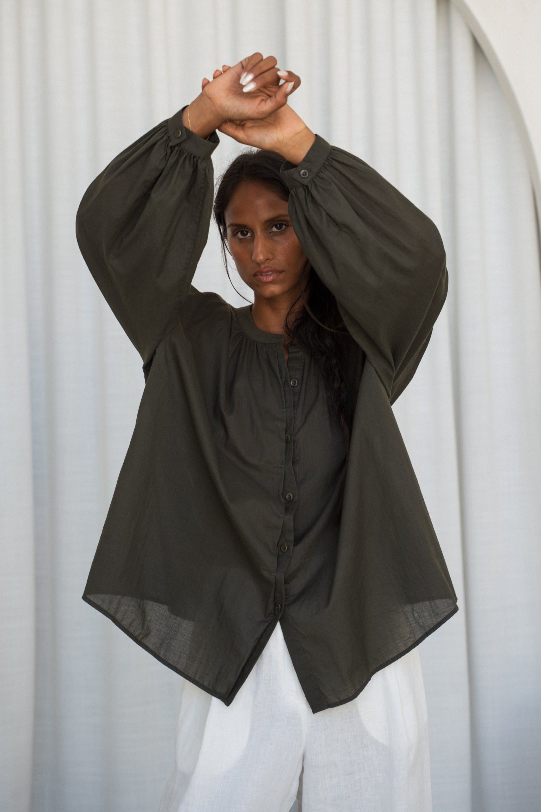 LILLY PILLY woman wearing a khaki Ember cotton voile shirt and ivory linen pants against  a white curtain background.