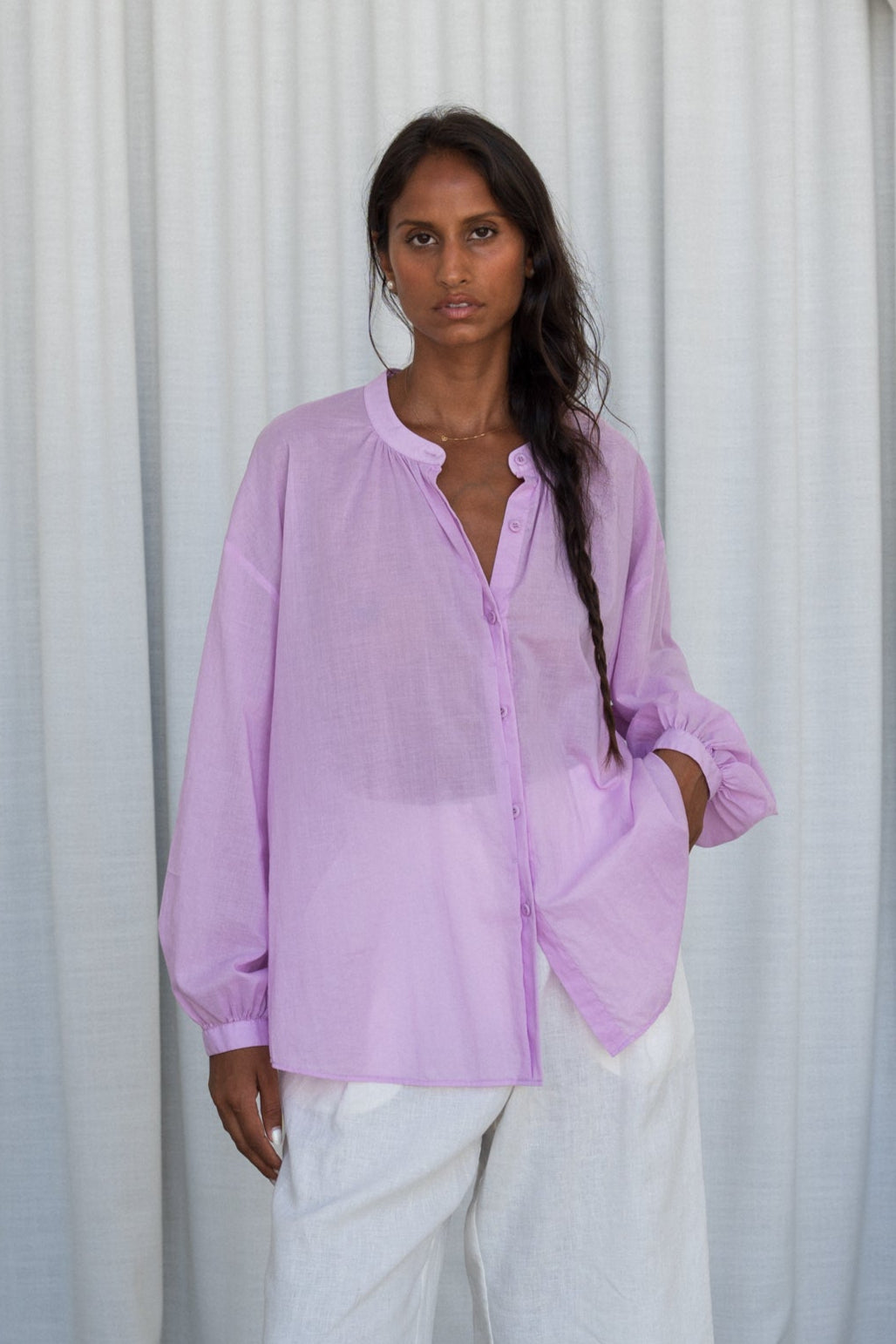 LILLY PILLY woman wearing a orchid Ember cotton voile shirt and white pants against a white curtain background showing front view
