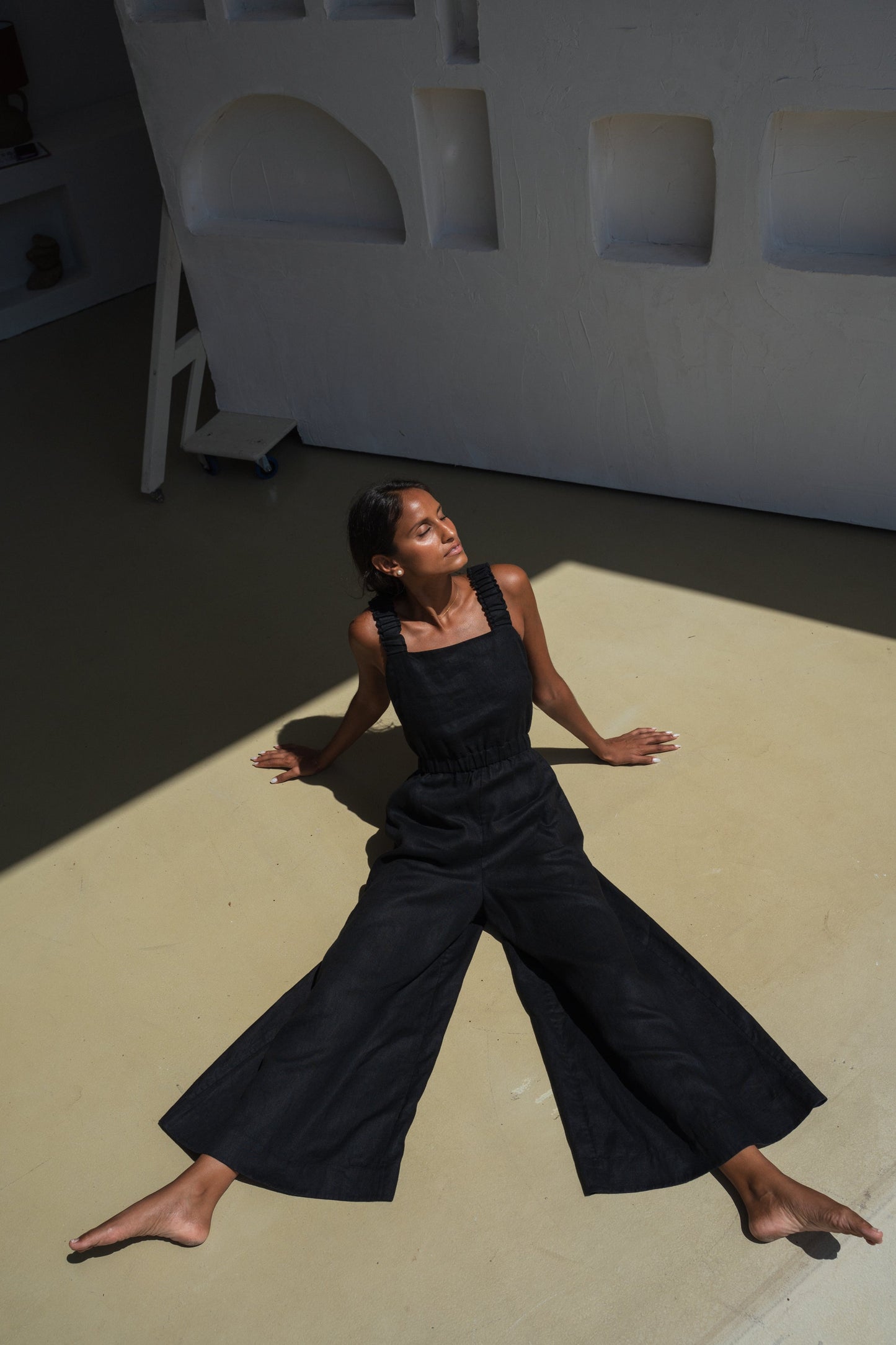 LILLY PILLY woman wearing a black Keely linen jumpsuit sitting in the sun with legs spread wide