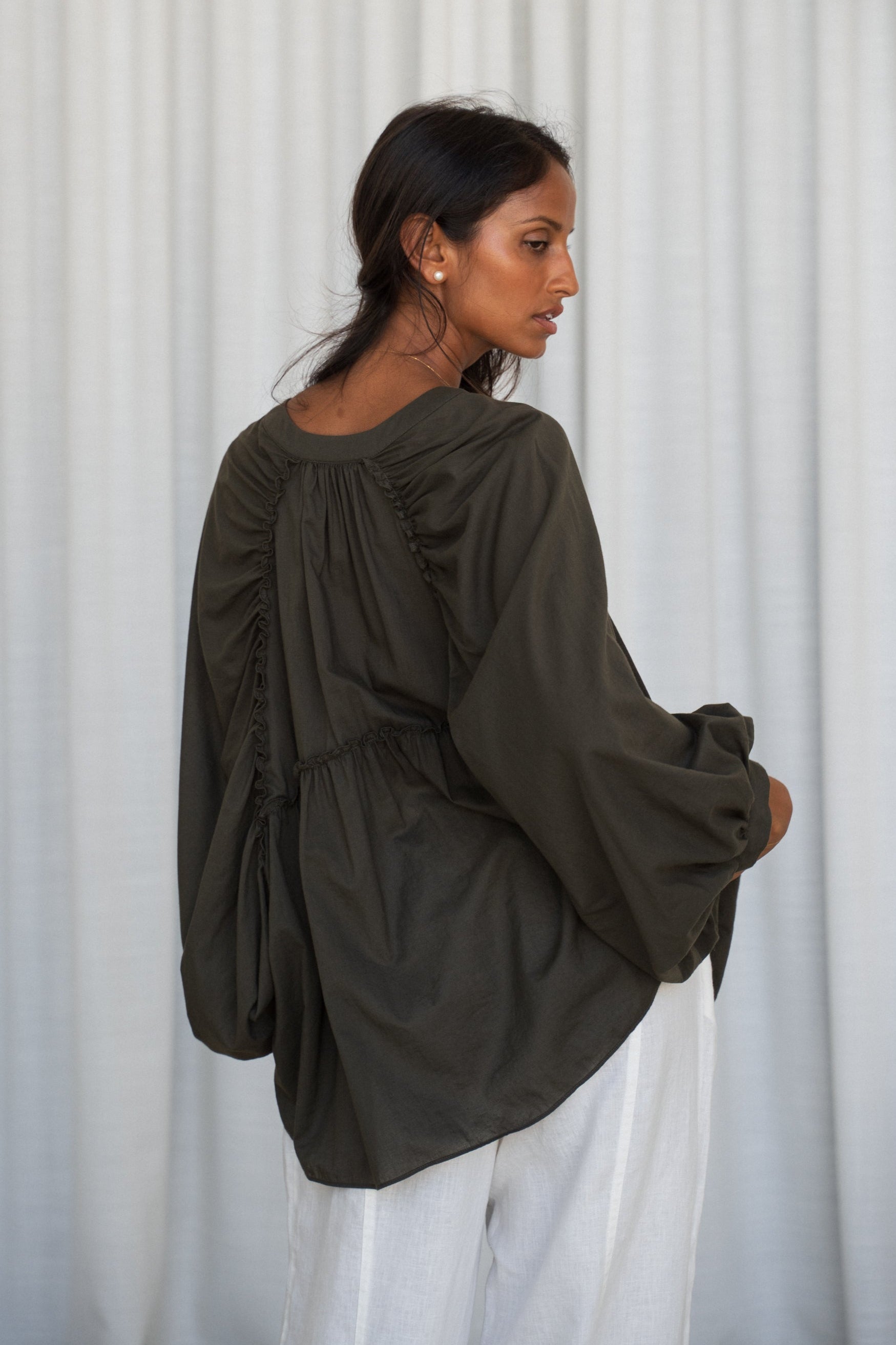LILLY PILLY woman wearing a khaki Thalia cotton voile shirt  blouse showing back view in front of a curtain background